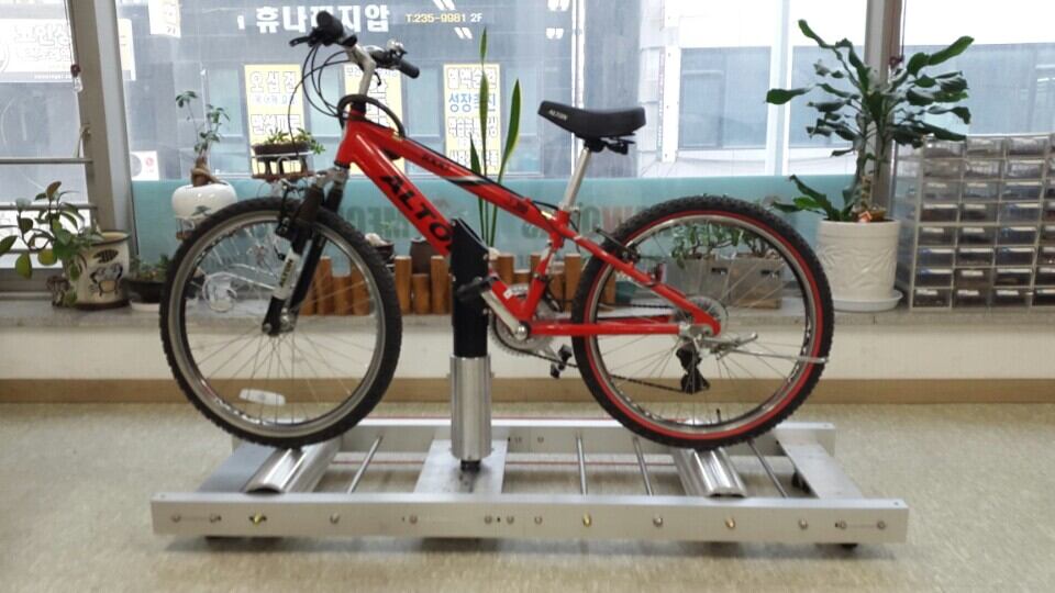 中央の支柱で赤い自転車を固定した、銀色のアルミ製ローラーフレームの初期Ultiracer試作機