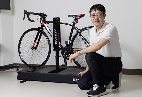 Joongsik Lee began building Ultiracer after a close call with a truck made riding outdoors hard RealDesignTech CEO Joongsik Lee kneeling beside a road bike mounted on an Ultiracer platform