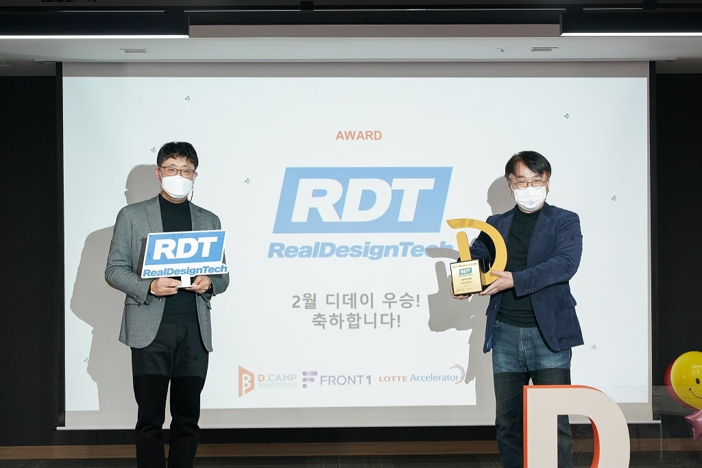 RealDesignTech gana el primer puesto en la D.DAY de febrero de 2021, organizada por d·camp con FRONT1 y Lotte Accelerator Dos representantes de RealDesignTech en el escenario con un cartel de RDT y el trofeo de la D.DAY, frente a una pantalla que felicita al ganador de febrero