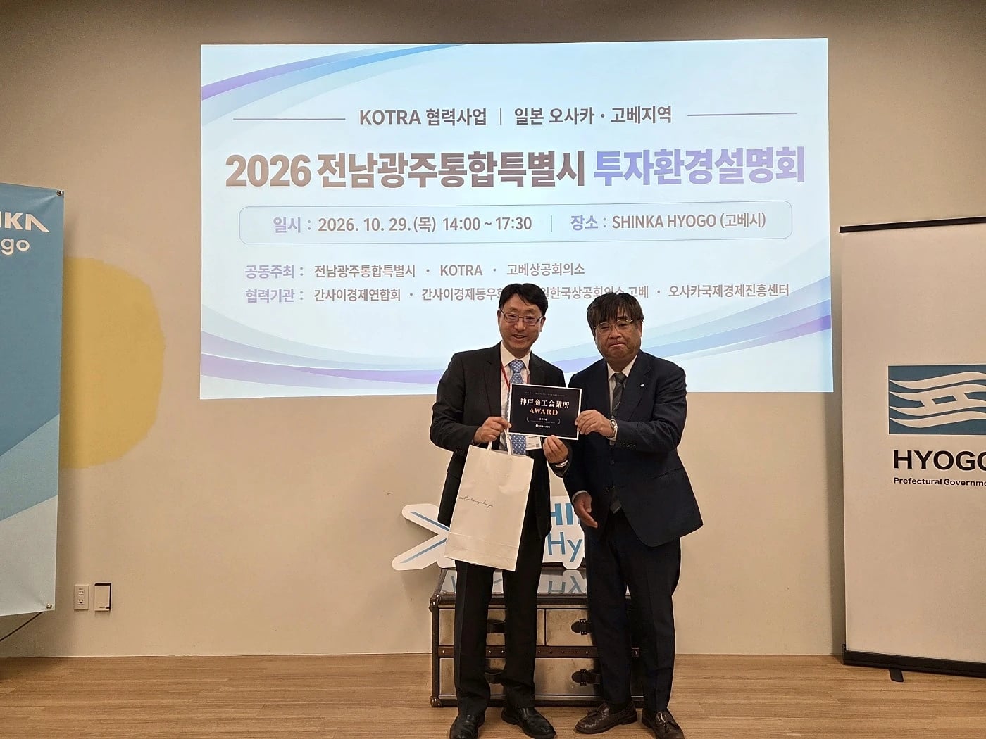 RealDesignTech CEO John Lee and a presenter holding the Kobe Chamber of Commerce and Industry Award card on stage at SHINKA Hyogo, Kobe, in front of a screen showing the event's Korean title, the 2026 Jeonnam-Gwangju Integrated Special City Investment Environment Briefing