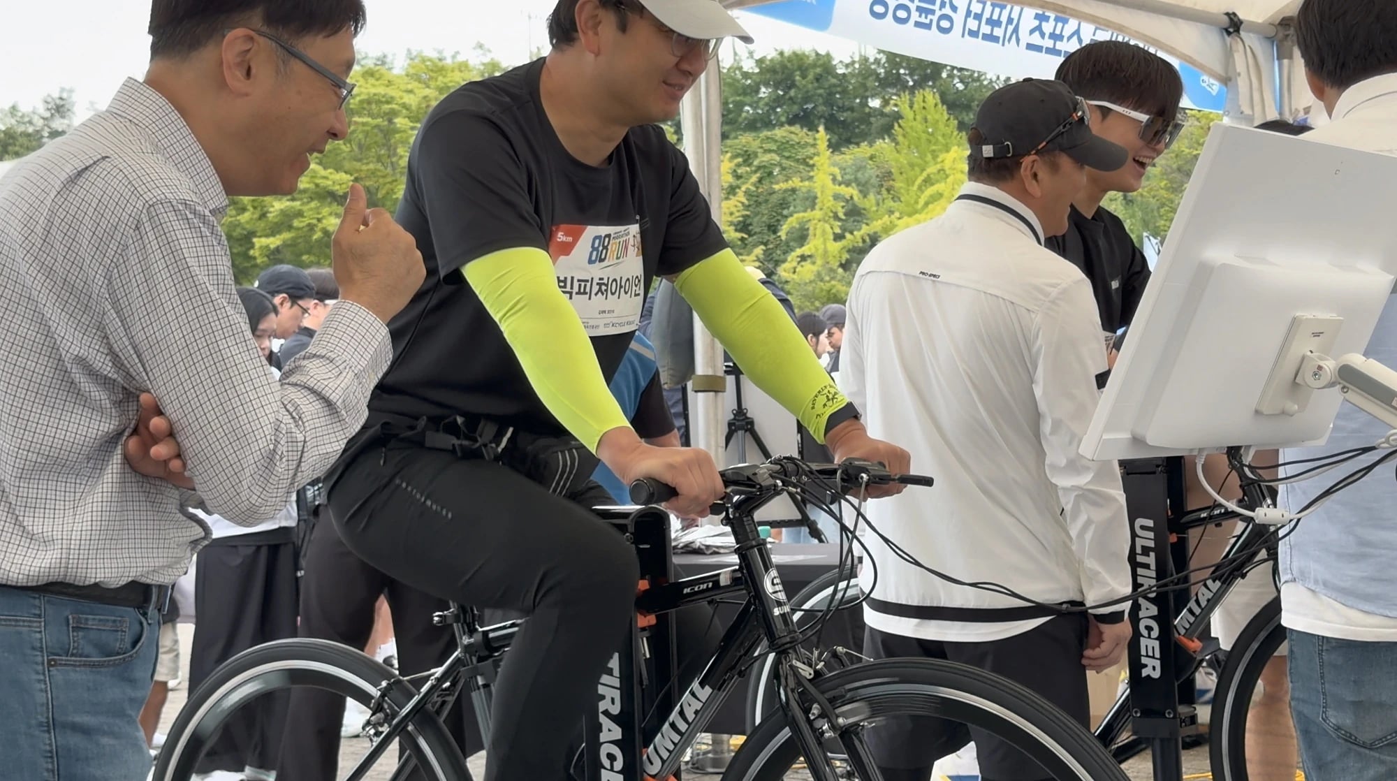 Ultiracer (얼티레이서) 자전거를 탄 5km 완주자 88RUN 5km 배번을 달고 연두색 팔토시를 낀 남성이 직원과 이야기하며 웃는 얼굴로 Ultiracer (얼티레이서) 자전거를 타는 모습