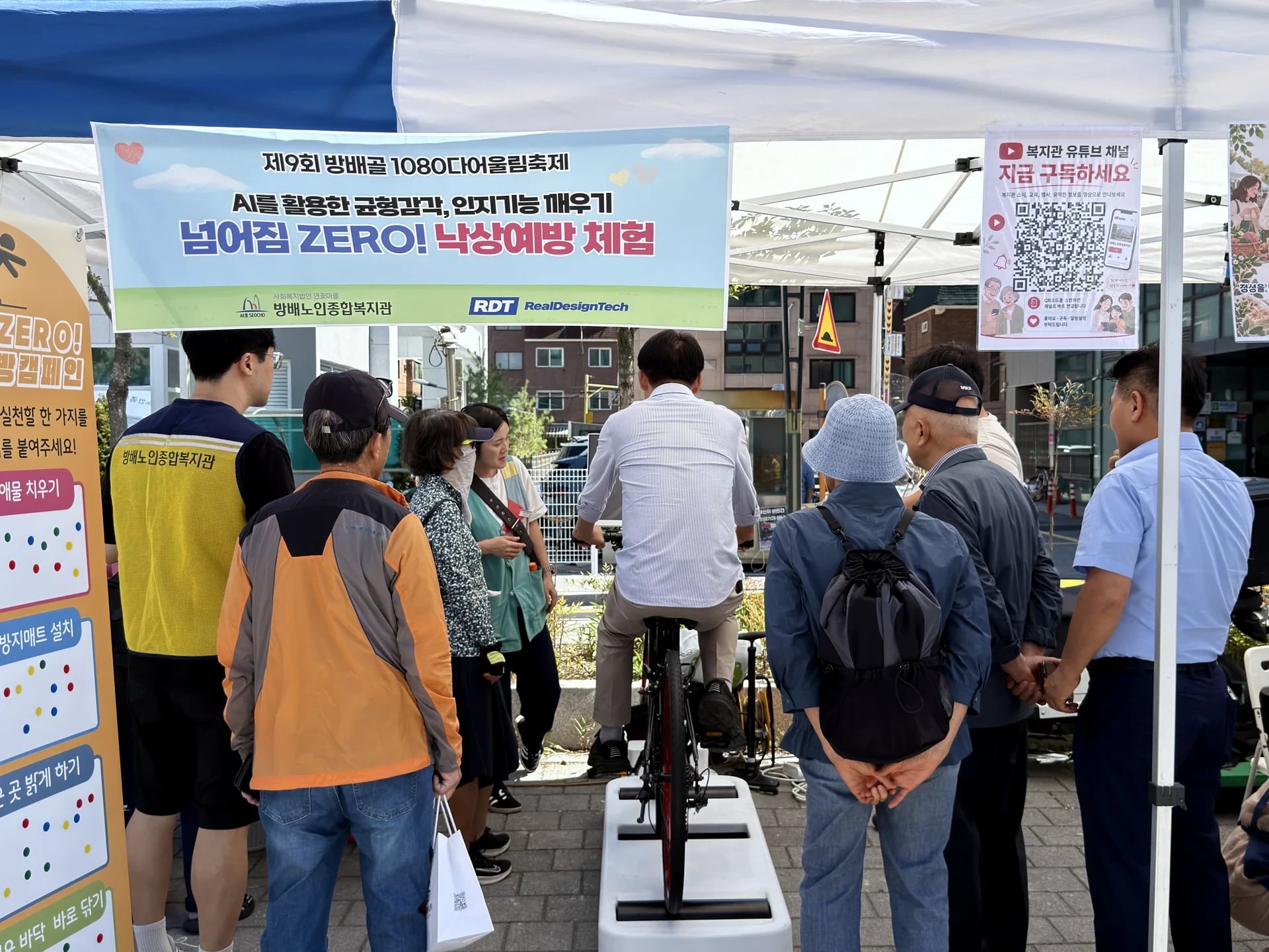 Older adults watching a senior ride the Ultiracer Kiosk under a banner reading 'Zero falls! Fall-prevention experience' at the 9th Bangbae-gol 1080 Festival, Seoul
