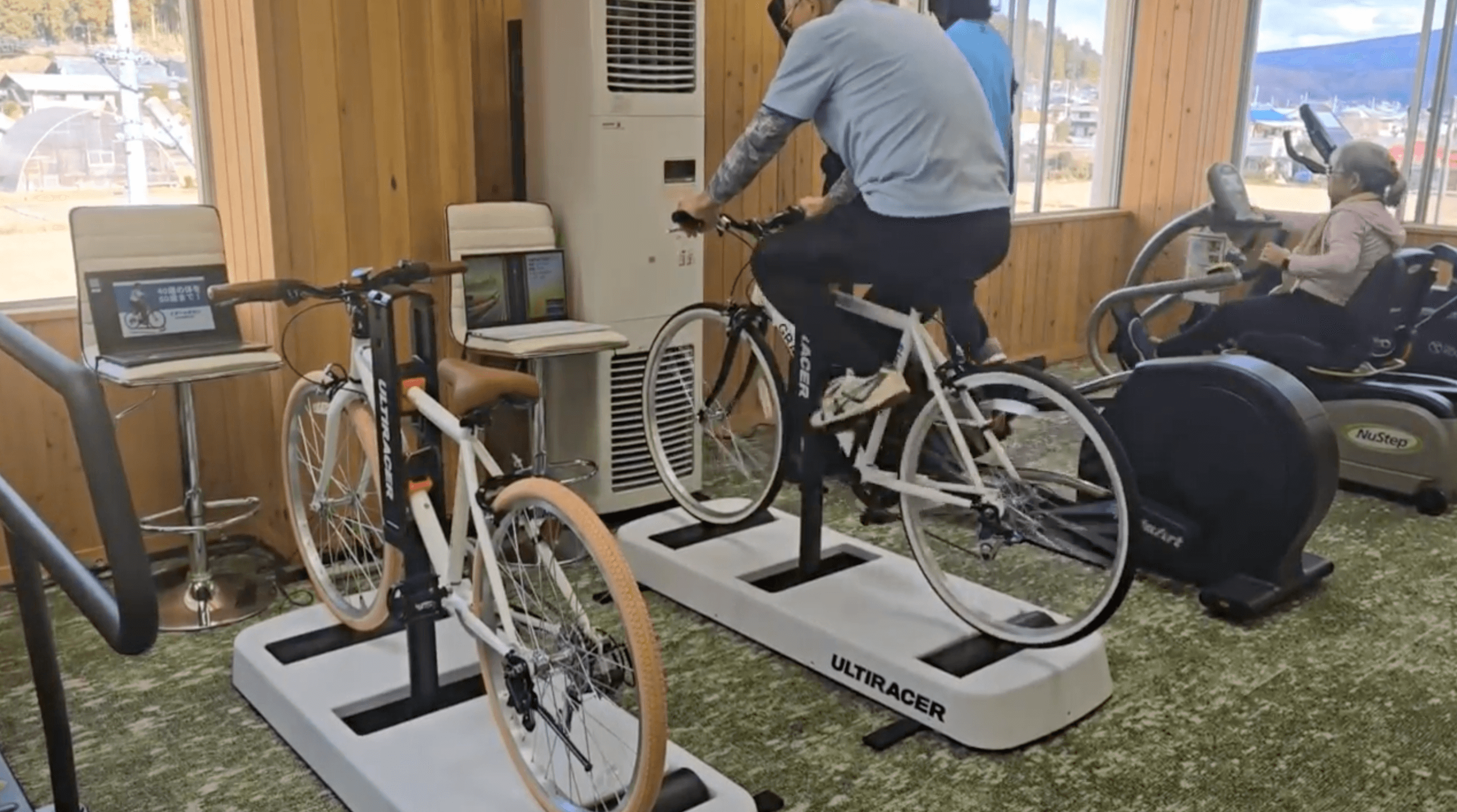 First rides in Nakanojo A man rides a bicycle on one of two Ultiracer platforms in a fitness room in Nakanojo, while an older woman exercises on a recumbent stepper nearby