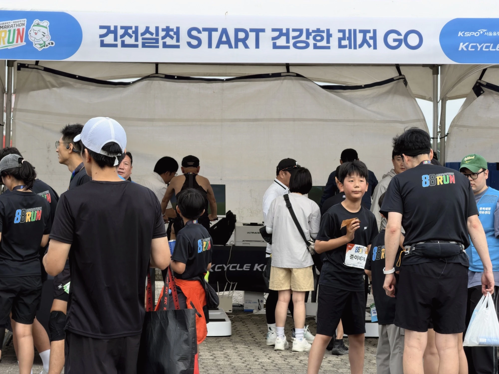 KCYCLE·KBOAT 부스 앞의 88RUN 러너들 88RUN 검은 티셔츠를 입고 배번을 단 러너들과 어린이들이 "건전실천 START 건강한 레저 GO" 배너가 걸린 흰 천막 앞에 모여 있는 모습