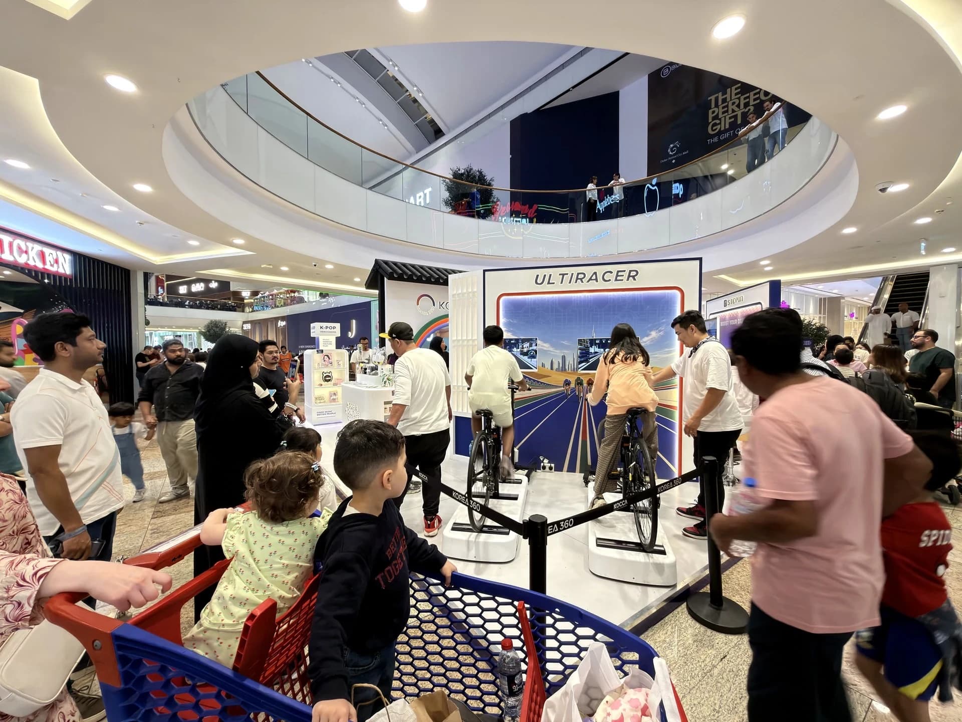 Crowd of families watching two visitors ride Ultiracer Kiosk systems at the KOREA 360 booth during Dubai K-Fest 2026, Dubai Festival City Mall