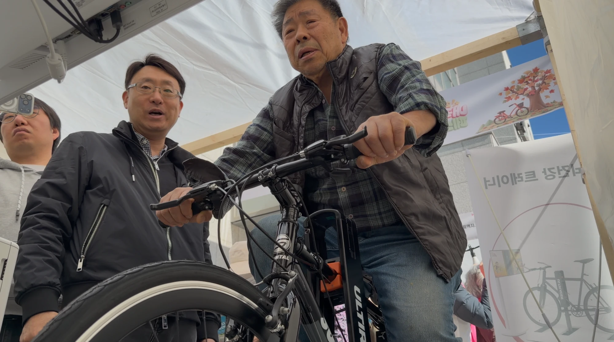A senior rider on Ultiracer Low-angle view of an older man in a plaid shirt and padded vest riding the Ultiracer bike and talking with a staff member standing beside him.