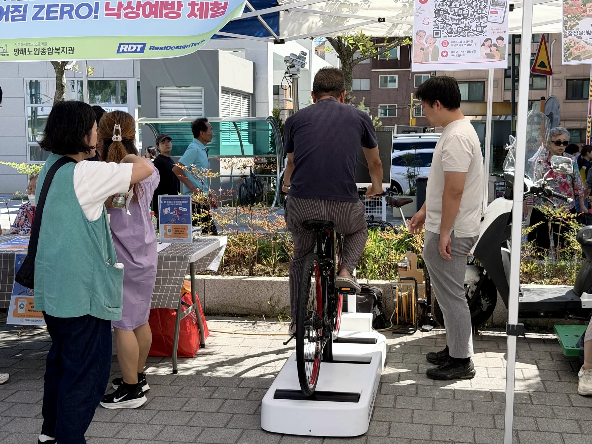 Ciclista mayor en el Ultiracer Kiosk del Centro de Bienestar para Mayores de Bangbae Un ciclista mayor pedalea en el Ultiracer Kiosk al aire libre junto al stand del Centro de Bienestar para Mayores de Bangbae mientras el personal y los vecinos observan