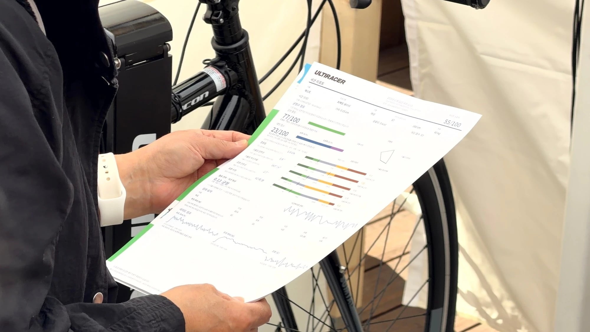 A printed Ultiracer balance report Close-up of a visitor holding a printed Ultiracer session report with green, blue and orange score bars and ride graphs, next to the bike.