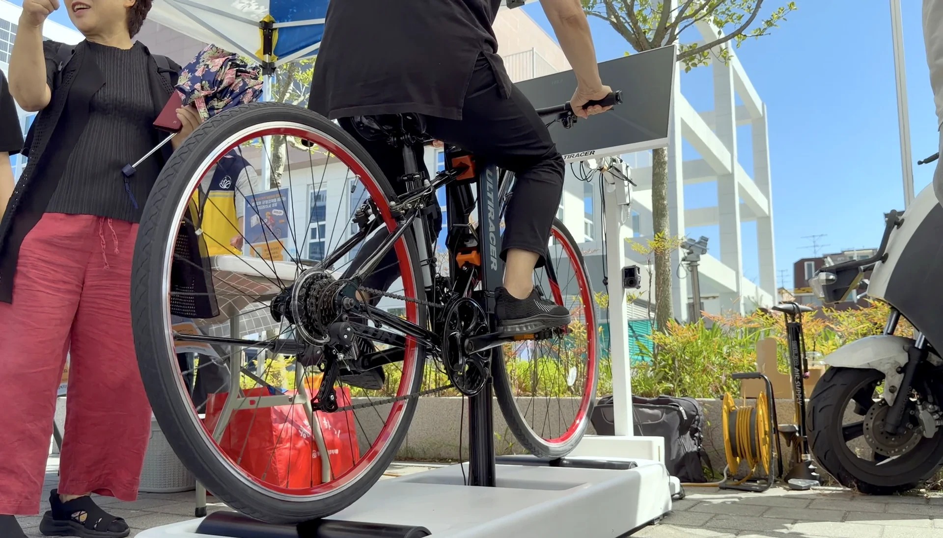 Plataforma de equilibrio del Ultiracer Kiosk en el Festival Bangbae-gol 1080 Vista a ras de suelo de los pies de un ciclista sobre los pedales del Ultiracer Kiosk, con ruedas de llanta roja sobre la plataforma blanca de equilibrio, en un festival comunitario al aire libre en Seúl