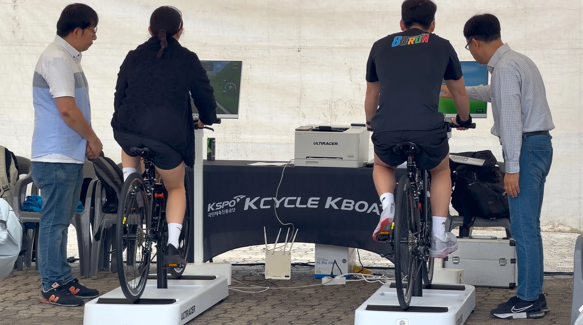KCYCLE·KBOAT 부스에서 라이더 곁을 지킨 직원 뒤에서 본 두 라이더가 화면 앞에서 Ultiracer (얼티레이서) 자전거 페달을 밟고, KSPO KCYCLE KBOAT 테이블 옆에 직원들이 서 있는 모습