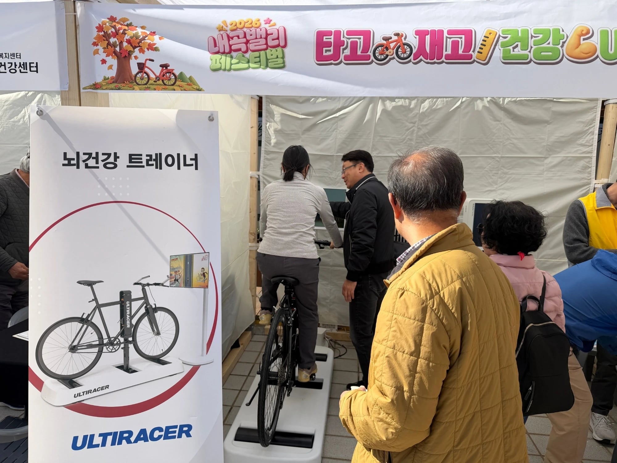A visitor rides the Ultiracer bike in the Neutinamu Shelter booth at the 2026 Naegok Valley Festival, guided by a staff member, while neighbours wait beside an Ultiracer banner.