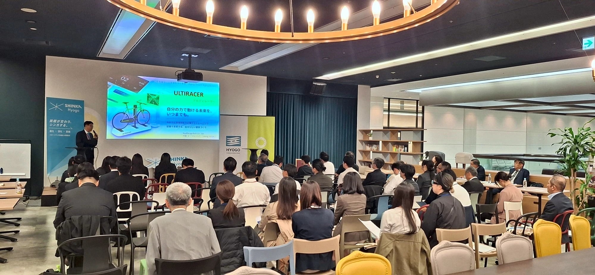 SHINKA Hyogo(神戸)でのUltiracerピッチ 神戸・SHINKA Hyogoで、Ultiracerのタイトルスライドの横の演台からJohn Leeが話し、満席の参加者が耳を傾けている様子。ステージ脇にSHINKA Hyogoと兵庫県のバナーが立っている