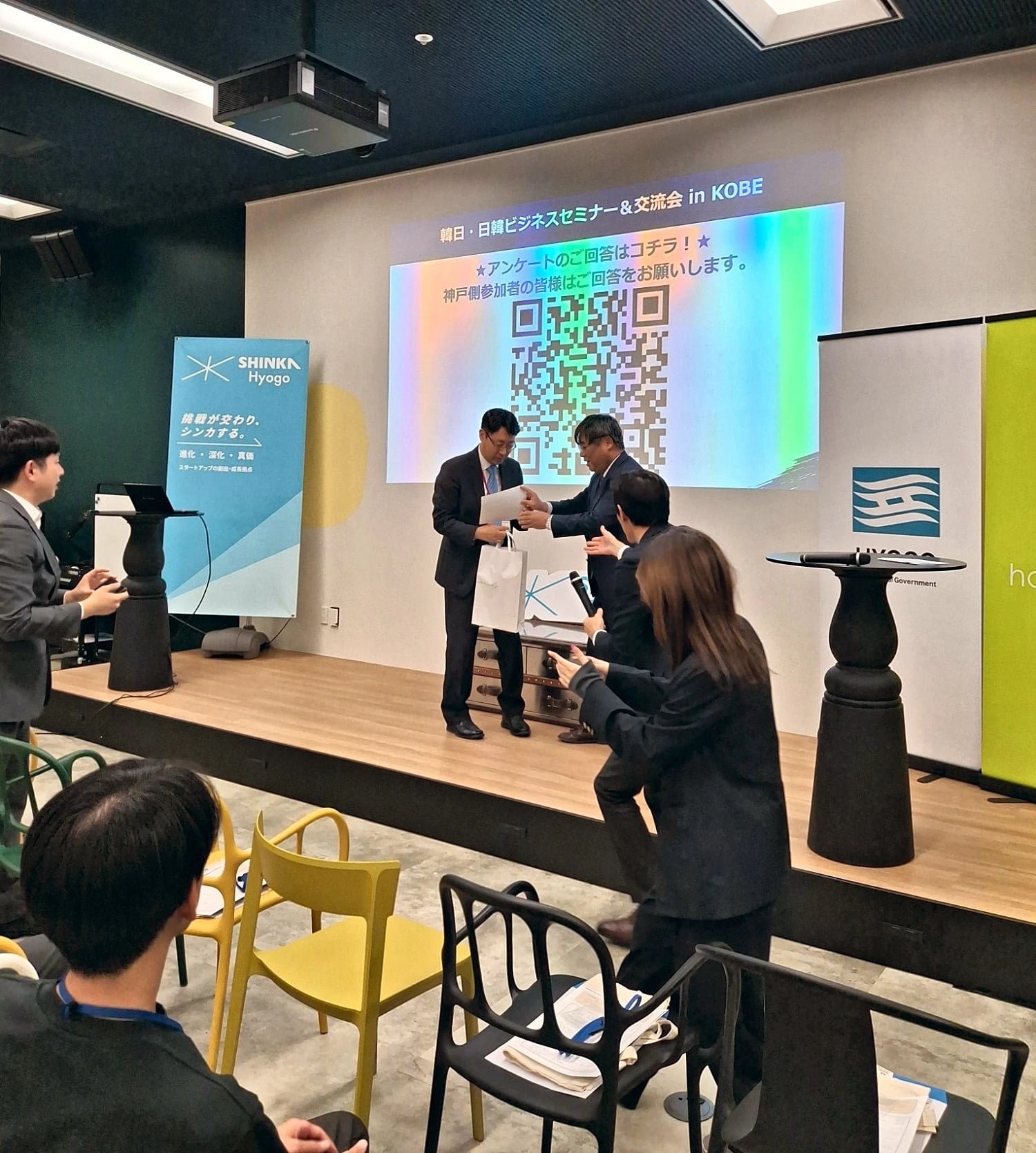 SHINKA Hyogoでの表彰 SHINKA Hyogoのステージで、John Leeがプレゼンターから賞を受け取り、ステージ脇の運営スタッフや参加者が拍手を送る様子