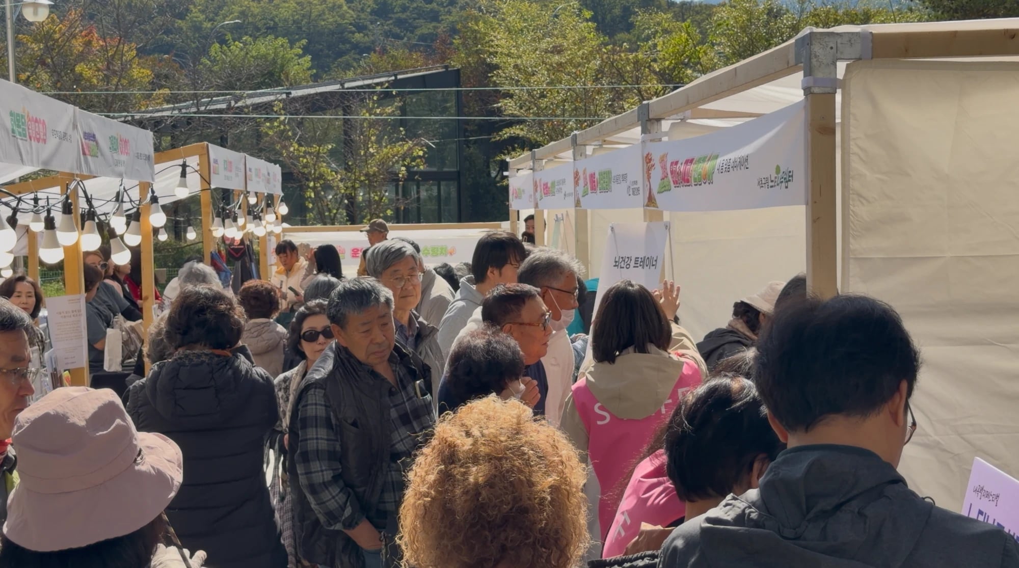 Booths at the 2026 Naegok Valley Festival Crowds of residents, many of them older adults, fill a lane between white festival booths on a sunny autumn day, with the Ultiracer booth banner visible on the right.