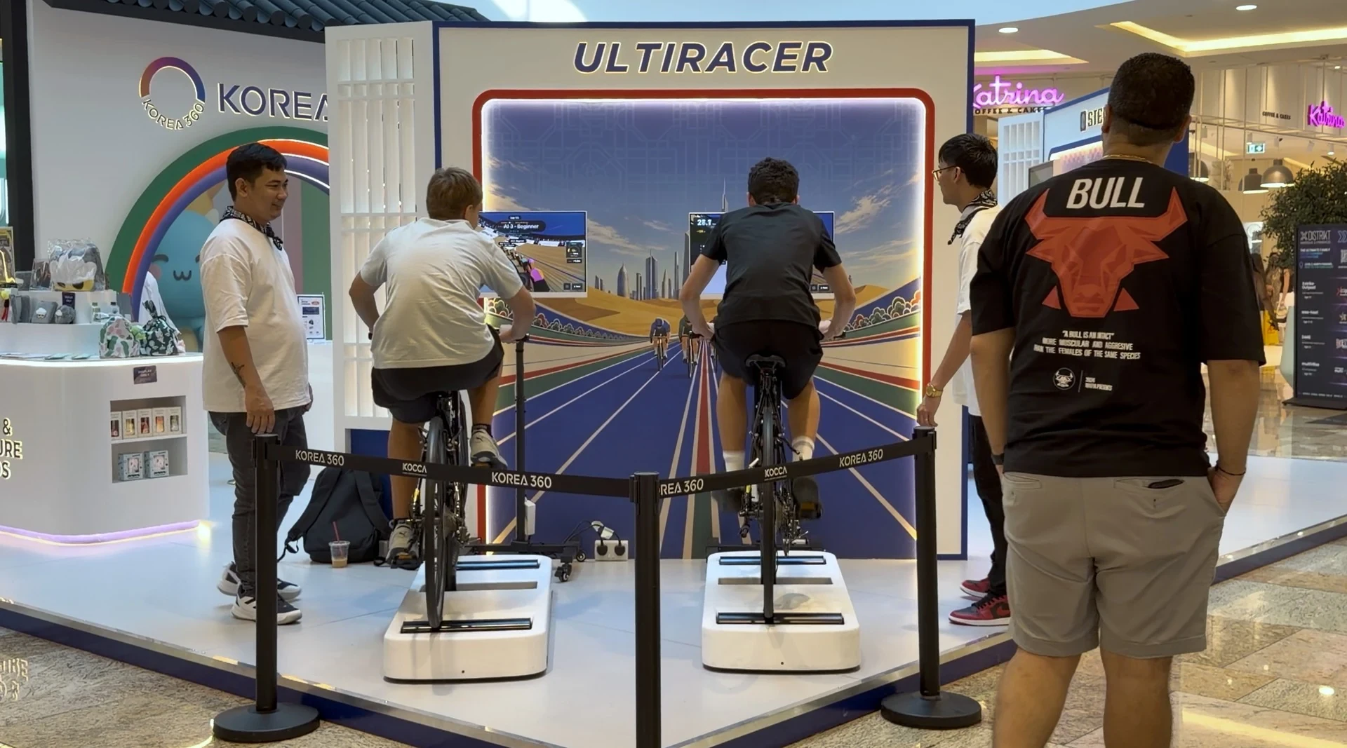 Ultiracerキオスクで対戦 ドバイ・フェスティバル・シティ・モールのKOREA 360ブースで、ULTIRACERの背景の前に並んだ2台のUltiracerキオスクでレースをする2人のライダー