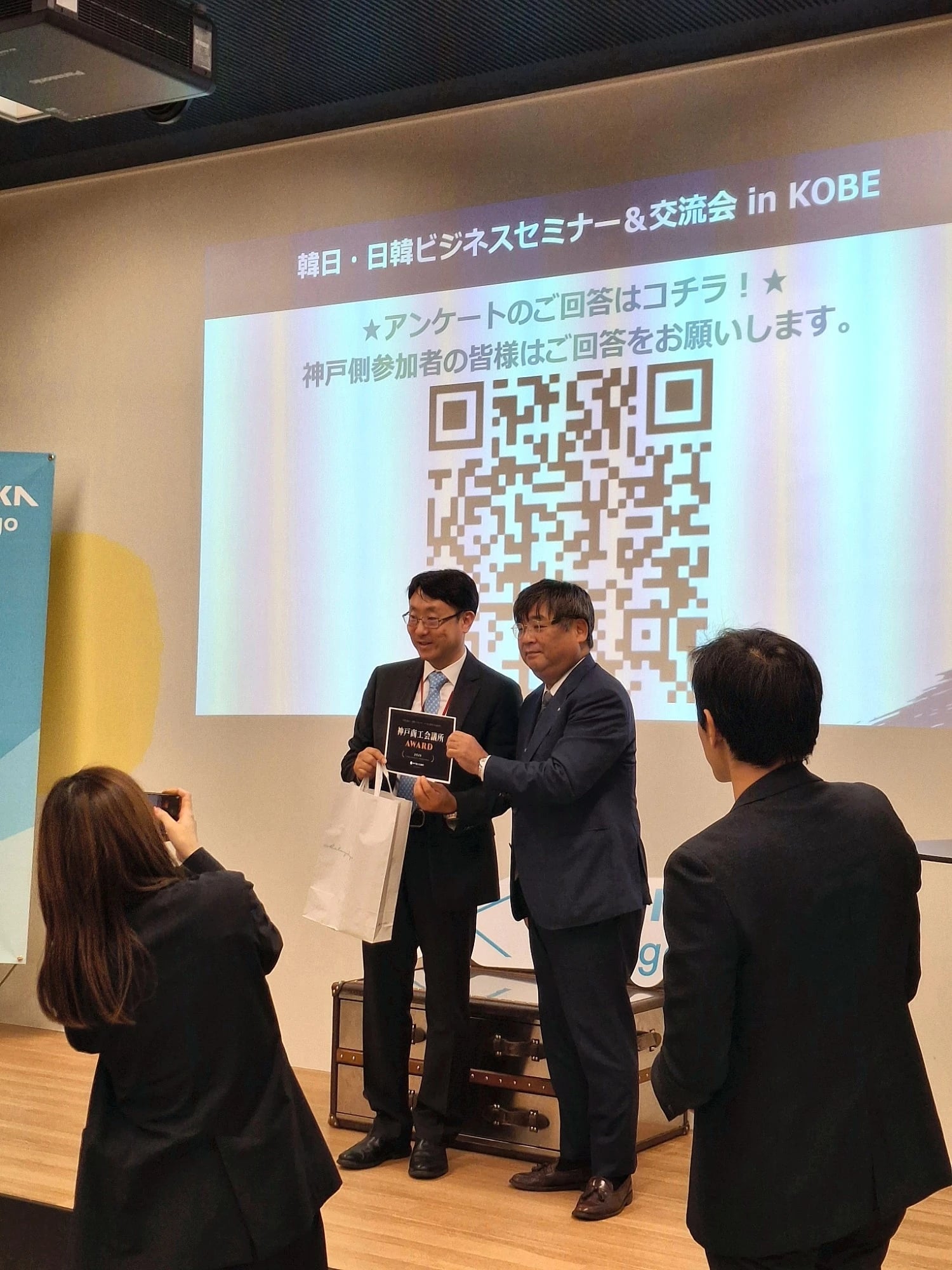 RealDesignTech CEO John Lee holding the Kobe Chamber of Commerce and Industry Award card with a presenter on stage at SHINKA Hyogo, Kobe, under the projected title of the Korea-Japan Business Seminar & Networking in KOBE