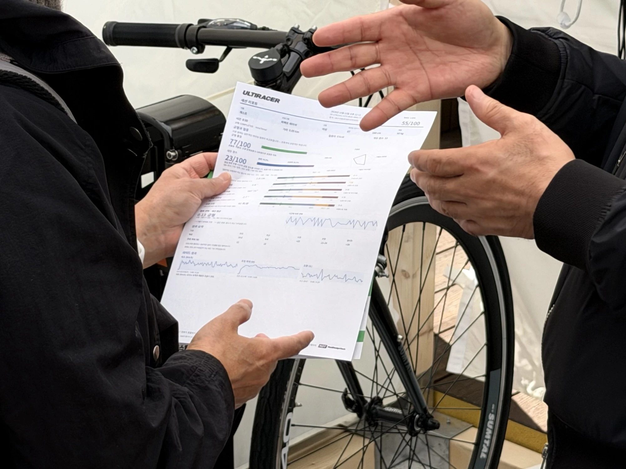 Walking a rider through the report A staff member gestures while explaining a printed Ultiracer session report that a rider holds in front of the bike.