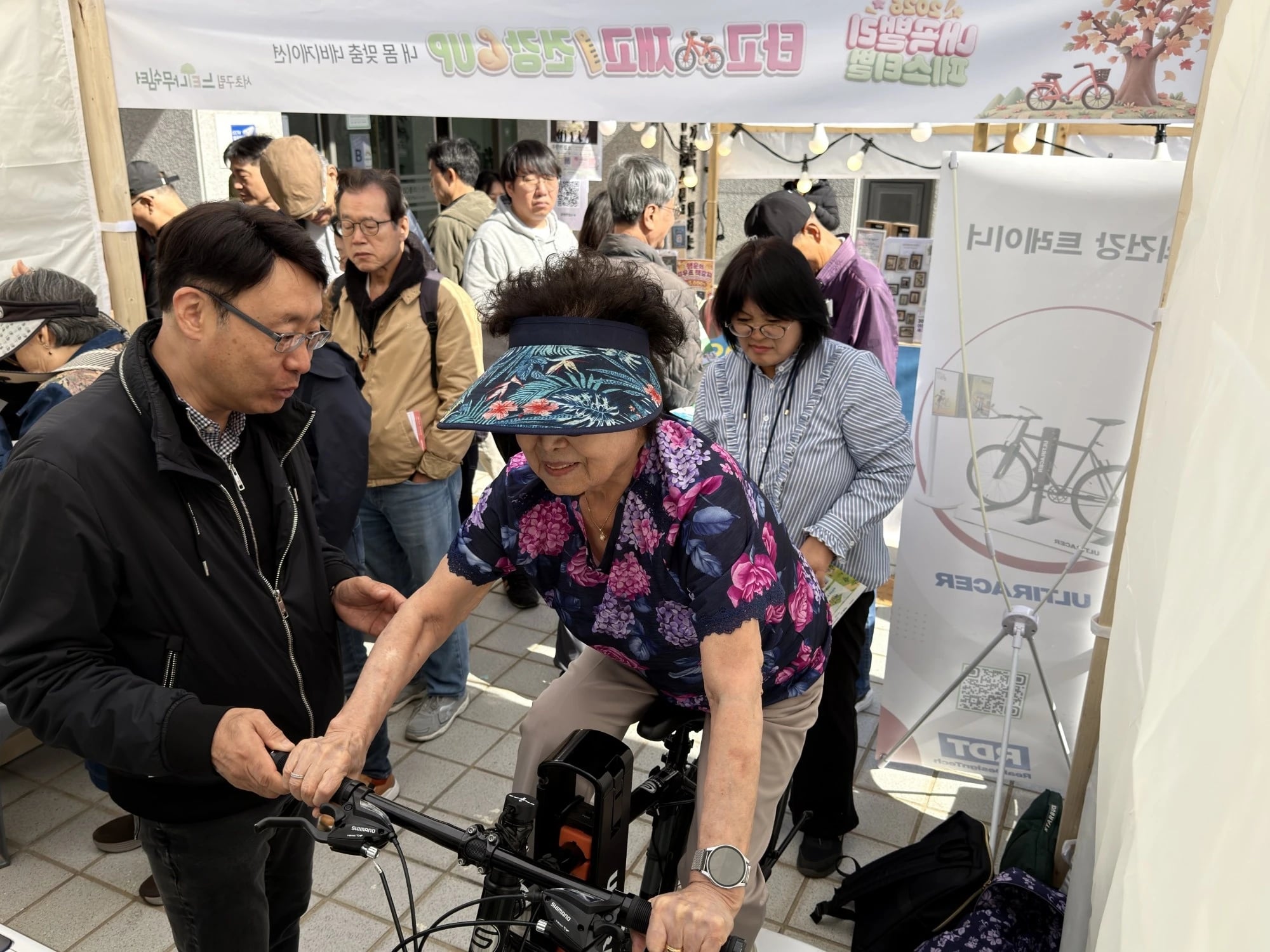 Settling in before the ride An older woman in a floral blouse and sun visor pedals the Ultiracer bike while a staff member guides her hand on the handlebar and visitors look on.