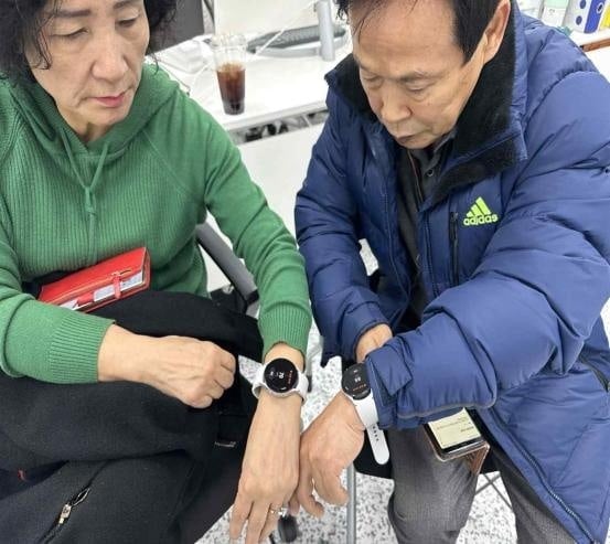 Two older participants seated side by side, each looking at the smartwatch on their wrist